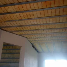 Poddasze w trakcie ocieplania wełną mineralną, widoczne metalowe profile konstrukcyjne, w tle okno z silnym światłem dziennym.