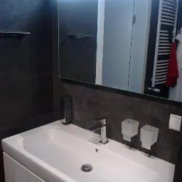 Nowoczesna umywalka z szafką w łazience wykończonej ciemną płytką, chromowana bateria i akcesoria, odbicie w lustrze ukazuje grzejnik drabinkowy.