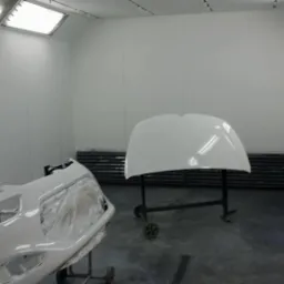 Biały zderzak samochodowy zabezpieczony folią i maską oraz biała maska samochodu na wózku w kabinie lakierniczej z jasnym oświetleniem.