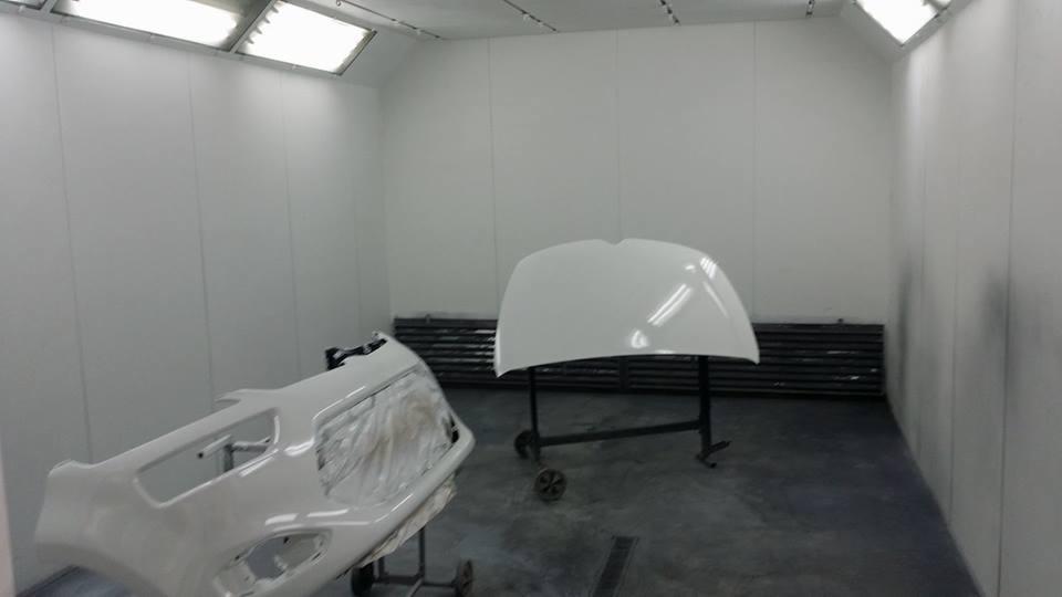 Biały zderzak samochodowy zabezpieczony folią i maską oraz biała maska samochodu na wózku w kabinie lakierniczej z jasnym oświetleniem.