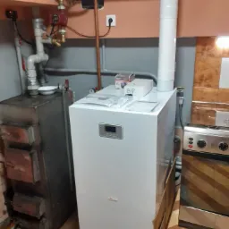 Nowoczesny, biały piec gazowy z elektronicznym wyświetlaczem, zainstalowany obok starszego pieca na paliwo stałe, widoczne rury odprowadzające spaliny i elementy instalacji grzewczej...