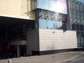 Fragment elewacji biurowca z jasnymi płytami elewacyjnymi i dużymi oknami, widoczny z poziomu chodnika, z refleksami słońca na szklanych powierzchniach.