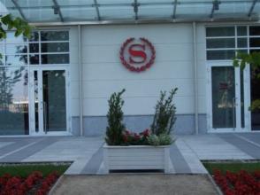 Elewacja budynku z symbolem 'S' w ozdobnym wieńcu, widoczne dwa wejścia z białymi ramami, dekoracyjny kwietnik z roślinami na pierwszym planie.