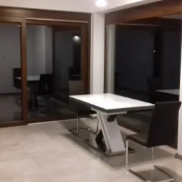 Jasne płytki podłogowe i białe ściany w nowoczesnym wnętrzu z dużymi przesuwnymi drzwiami i stołem z białym blatem i krzesłami.