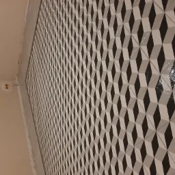 Ściana wyłożona błyszczącymi, trójwymiarowymi płytkami w odcieniach czerni, bieli i szarości, układającymi się w geometryczny wzór. Widoczne niedokończone wykończenia przy suficie i wystające...