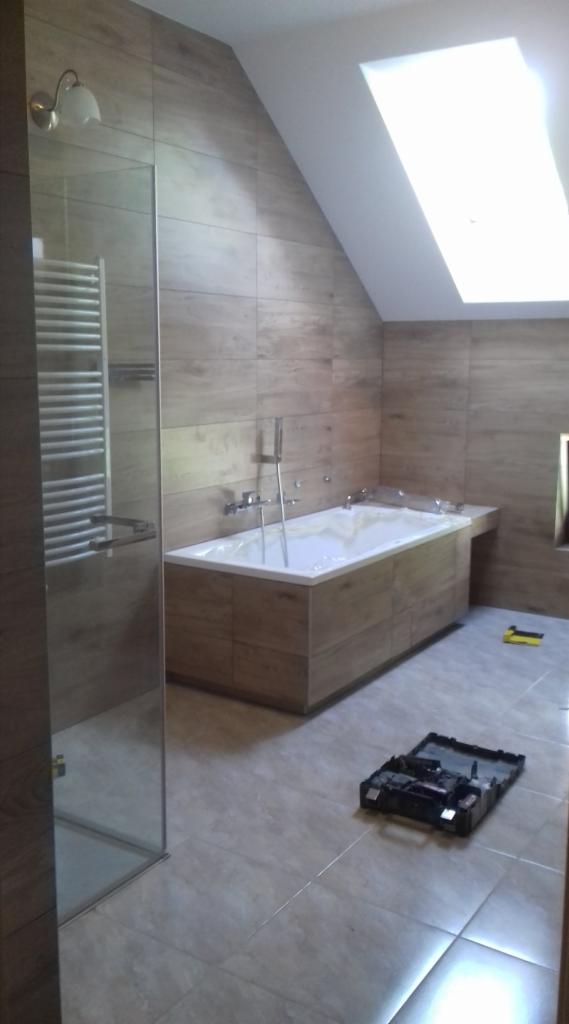 Nowoczesna łazienka z wanną obudowaną drewnopodobnymi panelami, szklaną kabiną prysznicową i oknem dachowym; na podłodze leżą narzędzia w otwartej skrzynce.