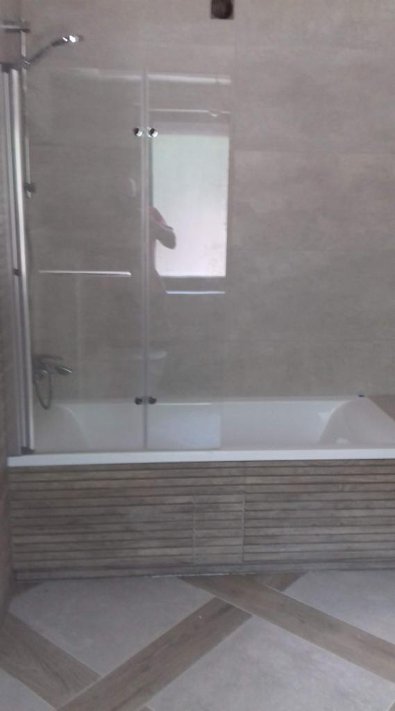 Zamontowana kabina prysznicowa z transparentnego szkła z chromowanymi elementami, widoczna biała wanna obudowana panelami imitującymi drewno, beżowe płytki na ścianach i podłodze ułożone we wzór...