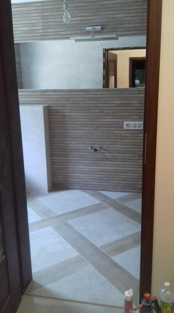 Widok na wykończoną łazienkę z elementami drewnopodobnymi na ścianach i geometryczną posadzką, widzianą zza uchylonych drzwi.