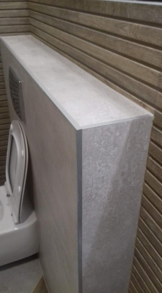 Obudowa stelaża podtynkowego WC wykończona płytkami imitującymi beton, z widoczną deską sedesową i fragmentem drewnopodobnej ściany w tle.