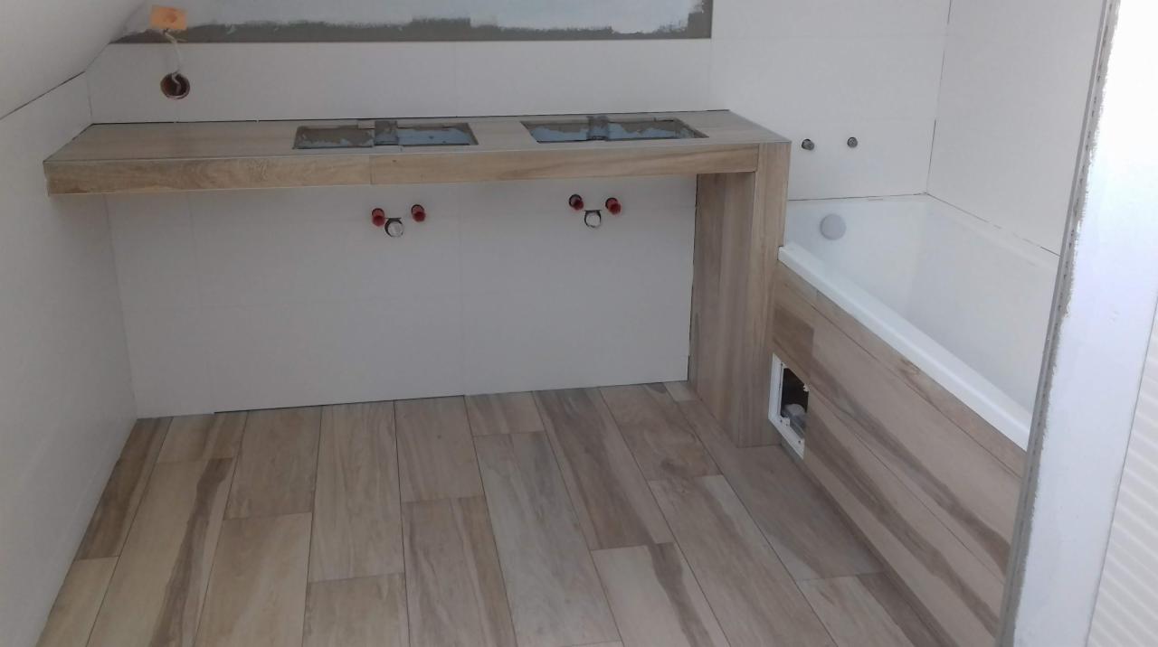 Wnętrze łazienki w trakcie prac wykończeniowych; widoczne przygotowane miejsce na dwie umywalki z doprowadzonymi instalacjami, obudowana wanna, płytki imitujące drewno na podłodze i ścianach.