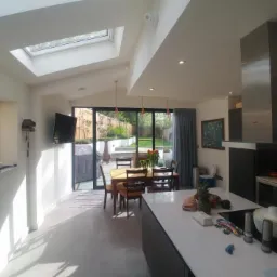 Panoramiczny widok na jasną kuchnię z wyspą, jadalnią i wyjściem na ogród, podkreślony świetlikiem dachowym i nowoczesnym okapem.