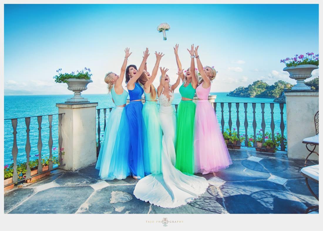 Panna młoda w białej sukni i druhny w kolorowych tiulowych spódnicach rzucają bukiet na tle morza i błękitnego nieba.