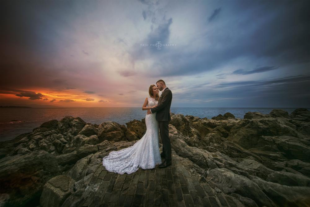 Para młoda w objęciach na skalistym wybrzeżu o zachodzie słońca, suknia ślubna z koronkowym trenem, widok na morze i dramatyczne niebo.