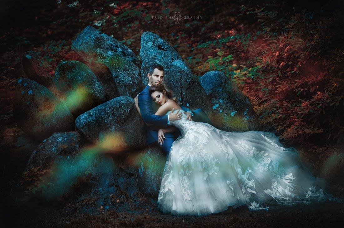 Para w strojach ślubnych pozuje na tle skalnej ściany w lesie, z efektem rozmycia i ciepłymi tonami.