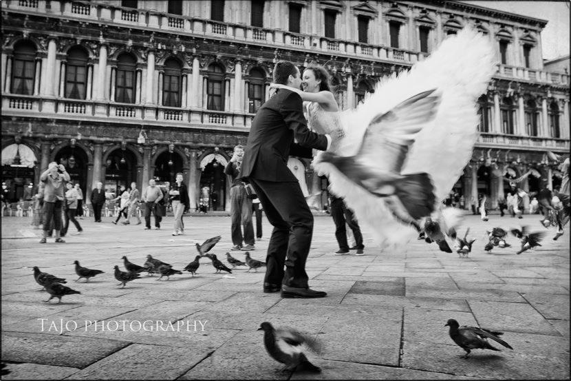 Czarno-biała fotografia pary młodej obejmującej się na tle historycznej architektury w Wenecji, z gołębiami w locie tworzącymi dynamiczny efekt.