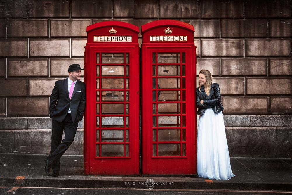 Para pozuje przy czerwonych budkach telefonicznych w Londynie: mężczyzna w garniturze z różowym krawatem i czapce z daszkiem, kobieta w skórzanej kurtce i długiej białej spódnicy.