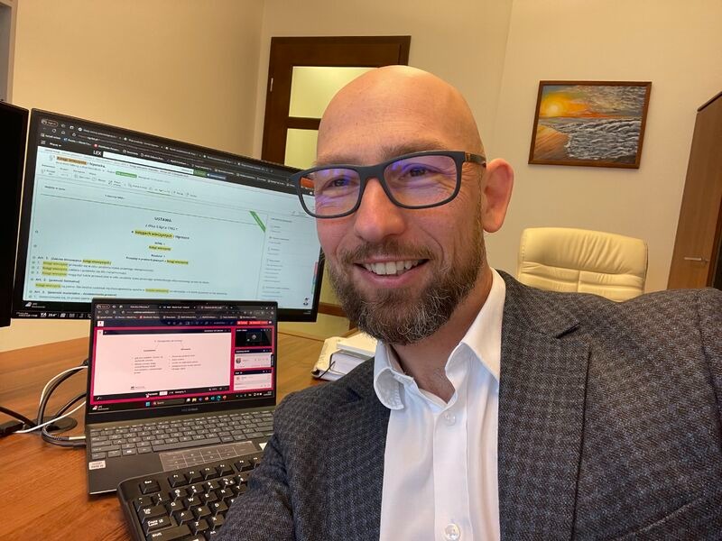 Uśmiechnięty mężczyzna w okularach i marynarce w biurze, z widocznymi ekranami komputera prezentującymi dokumenty prawne i wideokonferencję.