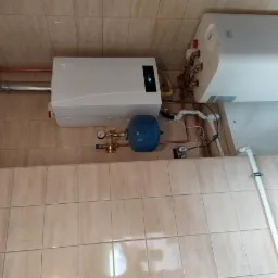 Biały piec gazowy kondensacyjny z podłączonym zasobnikiem ciepłej wody i niebieskim naczyniem przeponowym, zamontowane na ścianie wyłożonej beżowymi płytkami ceramicznymi. Widoczne rury...