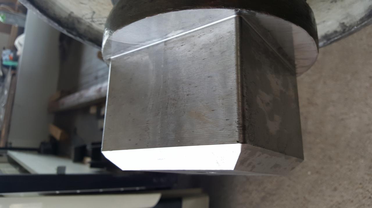 Szczegółowe ujęcie kwadratowego elementu metalowego, prawdopodobnie części maszyny, z widocznymi śladami obróbki i zużycia.