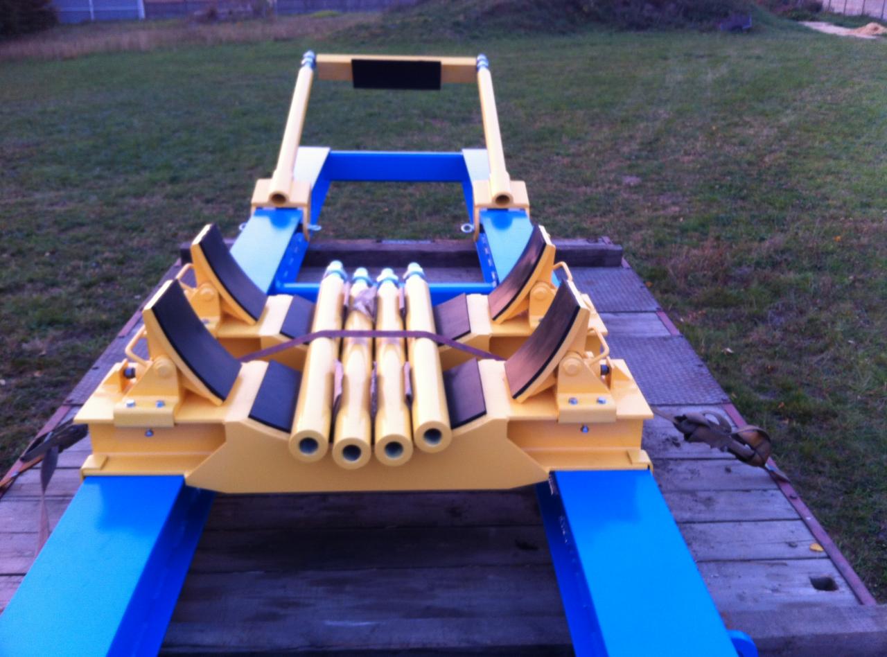 Żółto-niebieska konstrukcja stalowa na drewnianej platformie z sześcioma żółtymi rurami zabezpieczonymi pasami, widok z góry na tle trawnika.