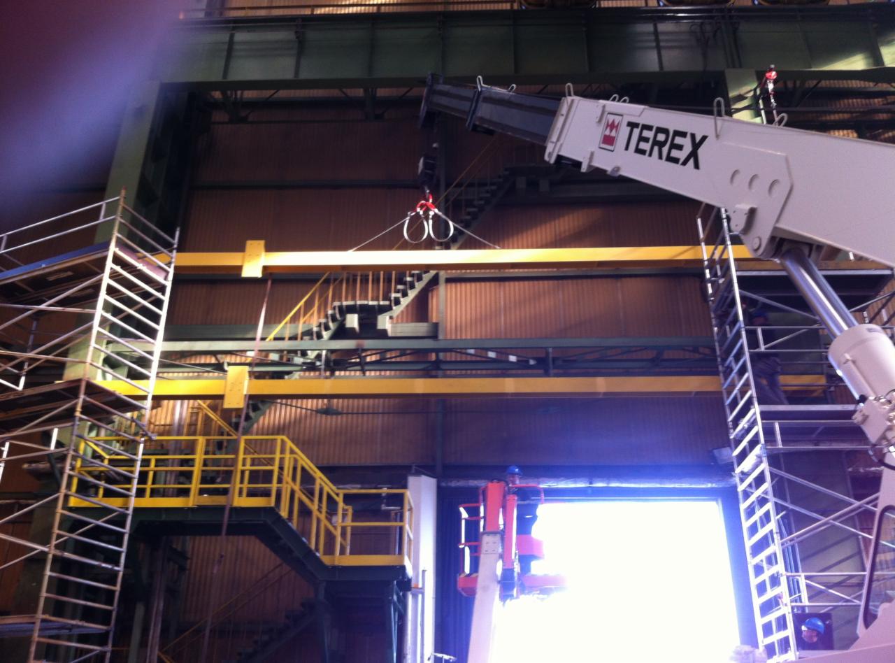 Montaż żółtej konstrukcji stalowej wewnątrz hali przemysłowej, widoczny dźwig Terex i rusztowania.