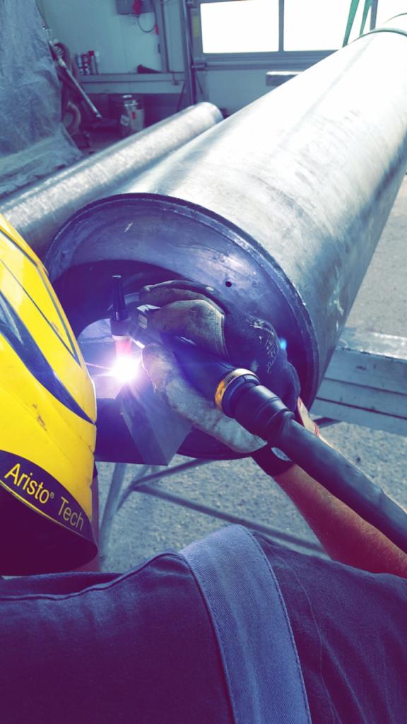 Spawanie rurociągu przez spawacza w masce Aristo Tech, widoczny jasny łuk spawalniczy i rękawice ochronne.