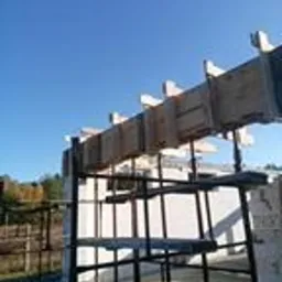 Szare bloczki fundamentowe z drewnianym szalunkiem pod wylewkę, wsparte na metalowej konstrukcji, na tle błękitnego nieba w słoneczny dzień.