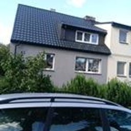 Robert Łyskawa - Dwukondygnacyjny dom z szarą elewacją, czarnym dachem i oknami z białymi ramami, częściowo zasłonięty przez zieleń i odbicie samochodu w dolnej części kadru.