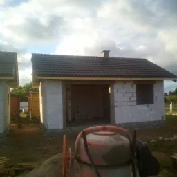 Niedokończony budynek z betoniarką na pierwszym planie. Widoczna konstrukcja dachu i ścian z pustaków. W tle pochmurne niebo.