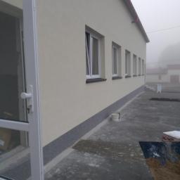 Robert Łyskawa - Świeżo pomalowana elewacja budynku z rzędem okien, widoczny fragment dachu i otwarte drzwi wejściowe, na pierwszym planie wiadro i taczka z piaskiem.