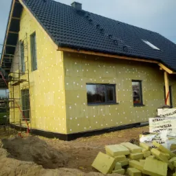 Dom jednorodzinny w trakcie ocieplania elewacji wełną mineralną, widoczne rusztowanie, stos wełny izolacyjnej i pryzma piasku.