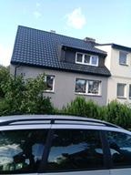 Dwukondygnacyjny dom z szarą elewacją, czarnym dachem i oknami z białymi ramami, częściowo zasłonięty przez zieleń i odbicie samochodu w dolnej części kadru.