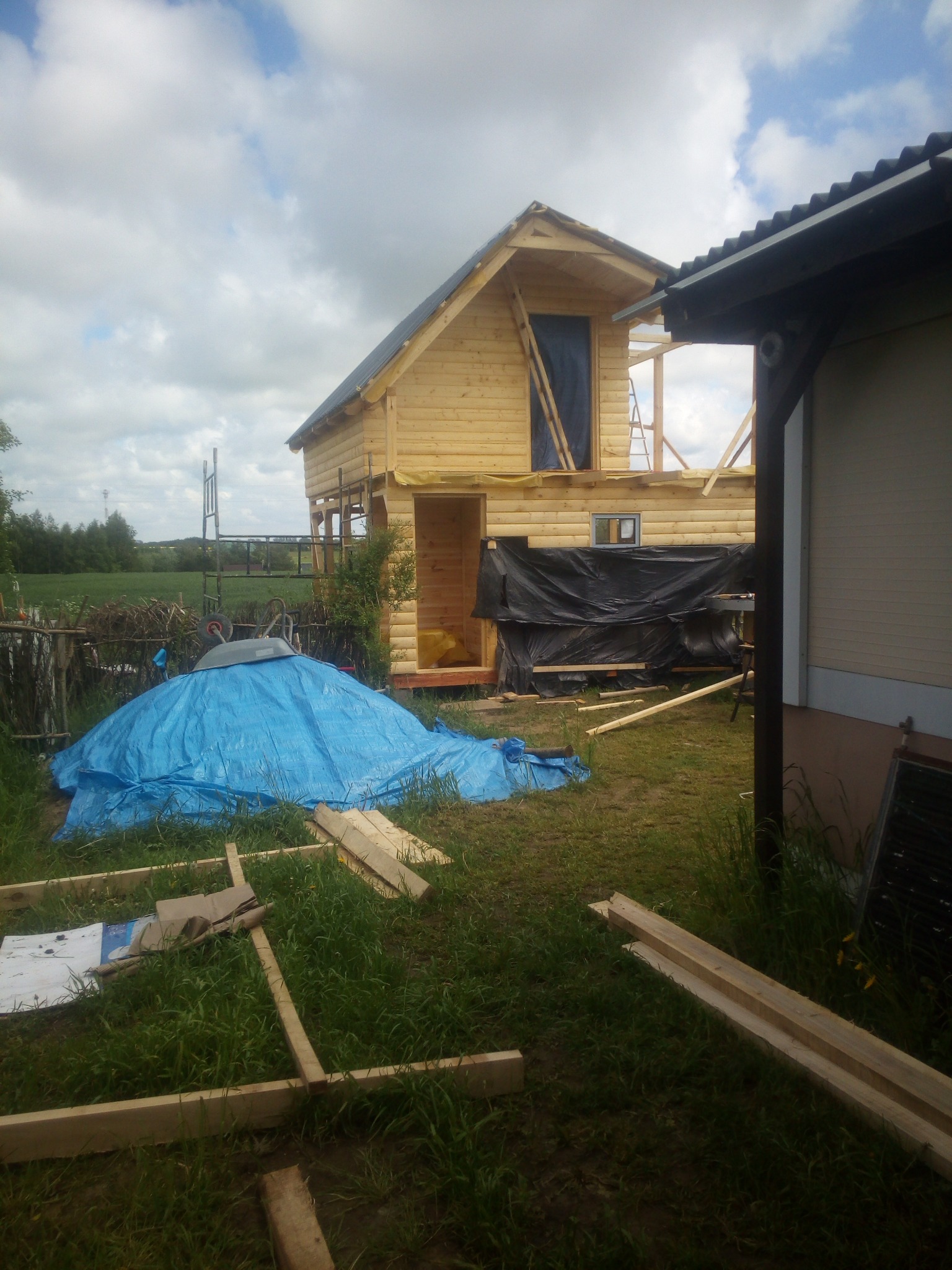 Surowy drewniany dom w trakcie budowy, z widocznymi elementami konstrukcyjnymi i zabezpieczeniami, na tle zielonego krajobrazu i pochmurnego nieba.