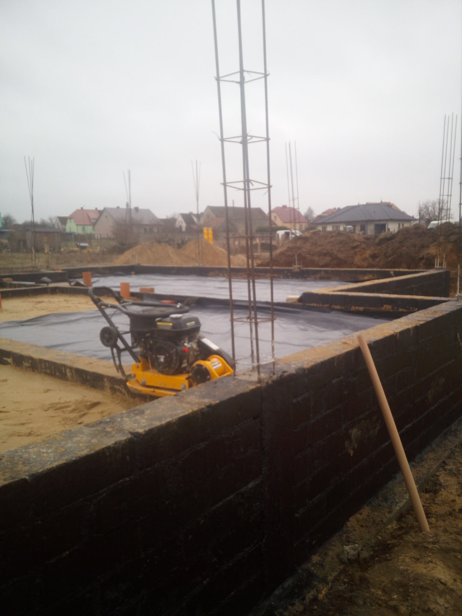 Fundamenty domu w trakcie budowy: zaizolowane ściany, folia hydroizolacyjna, zagęszczarka obok. W tle zabudowa jednorodzinna i szkielety zbrojenia.