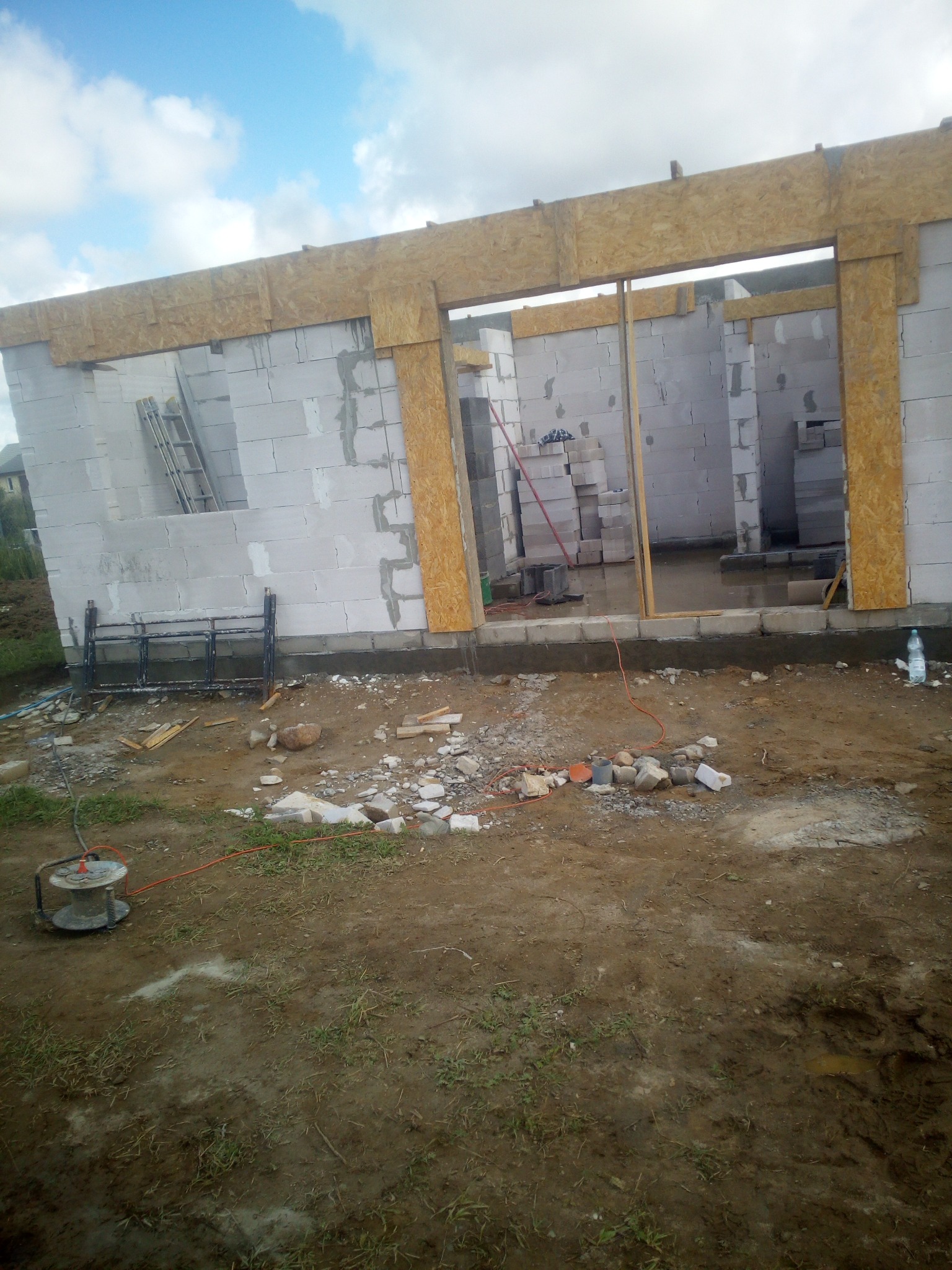 Wznoszenie ścian domu z betonu komórkowego, widoczne nadproża z płyty OSB, teren budowy z gruzem i narzędziami, nieukończony budynek w trakcie budowy.