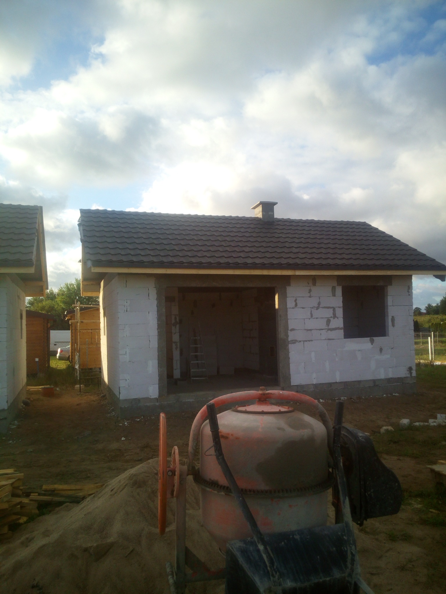 Niedokończony budynek z betoniarką na pierwszym planie. Widoczna konstrukcja dachu i ścian z pustaków. W tle pochmurne niebo.