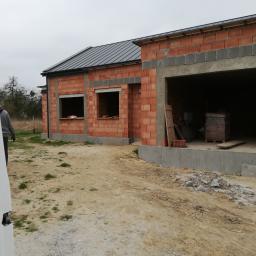 AGBUD KAROL MANIA - Surowy stan domu z czerwonej cegły z szarym dachem, widoczne otwory okienne i garaż, teren wokół domu niezagospodarowany.