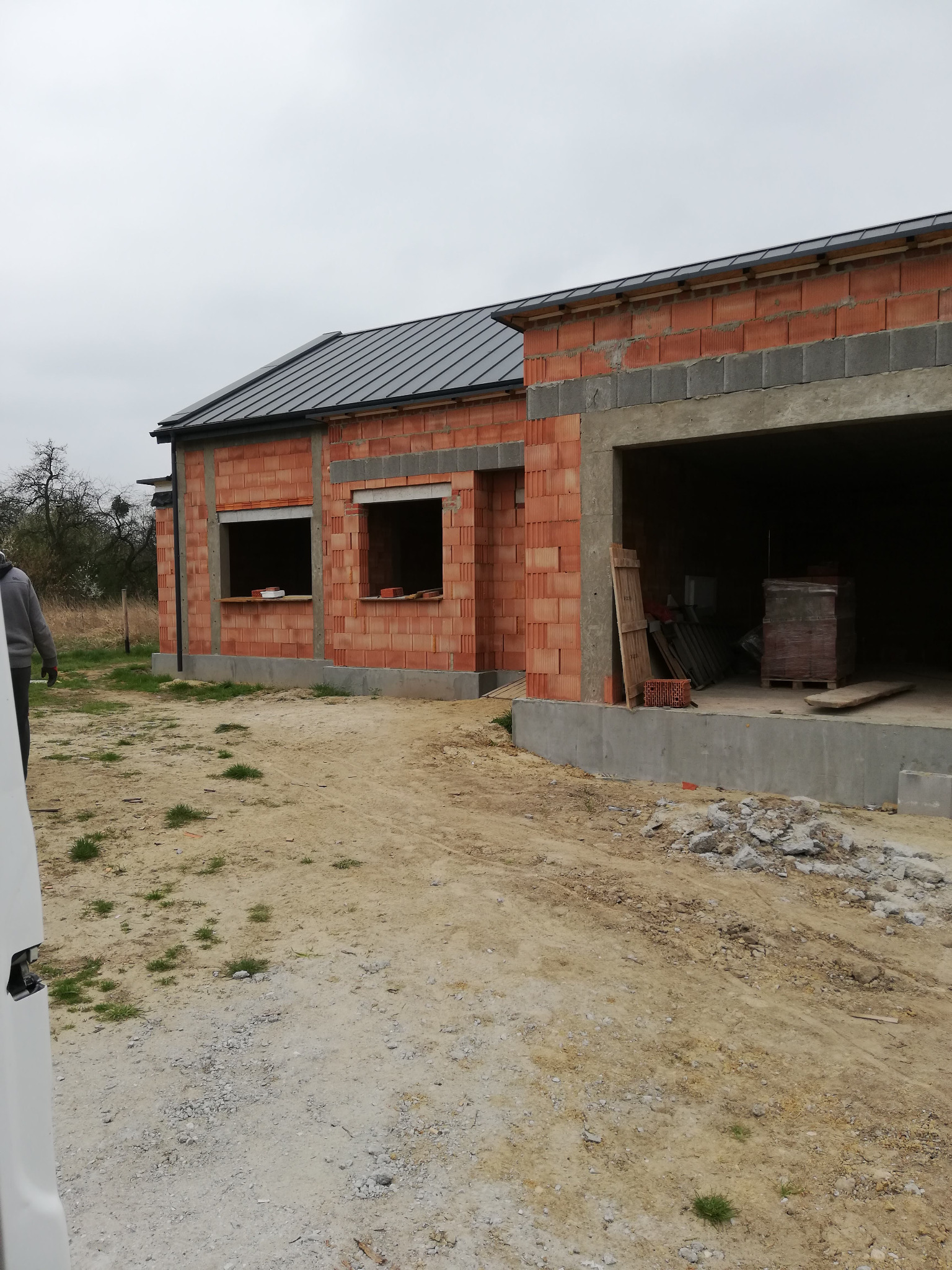 Surowy stan domu z czerwonej cegły z szarym dachem, widoczne otwory okienne i garaż, teren wokół domu niezagospodarowany.