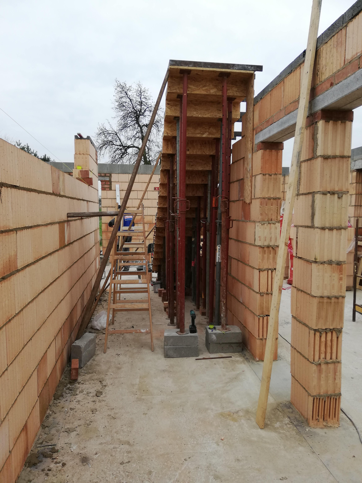 Wnętrze budowanego domu z czerwonej cegły ceramicznej, widoczne podpory stropowe, drabina i narzędzia budowlane.