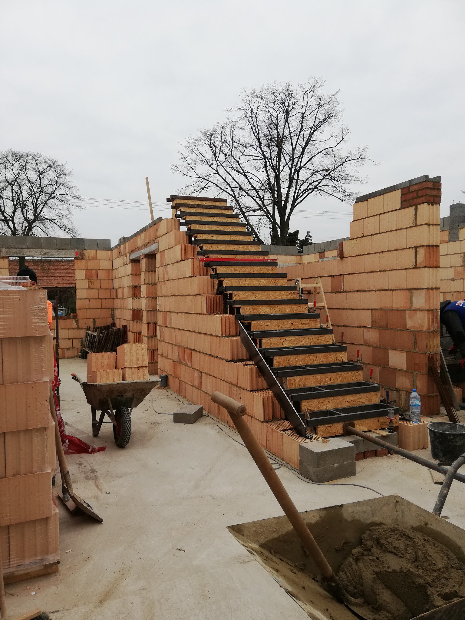 Wnętrze domu w trakcie budowy z czerwonej cegły, widoczne schody z metalową konstrukcją i drewnianymi stopniami, taczka z zaprawą i narzędzia budowlane.