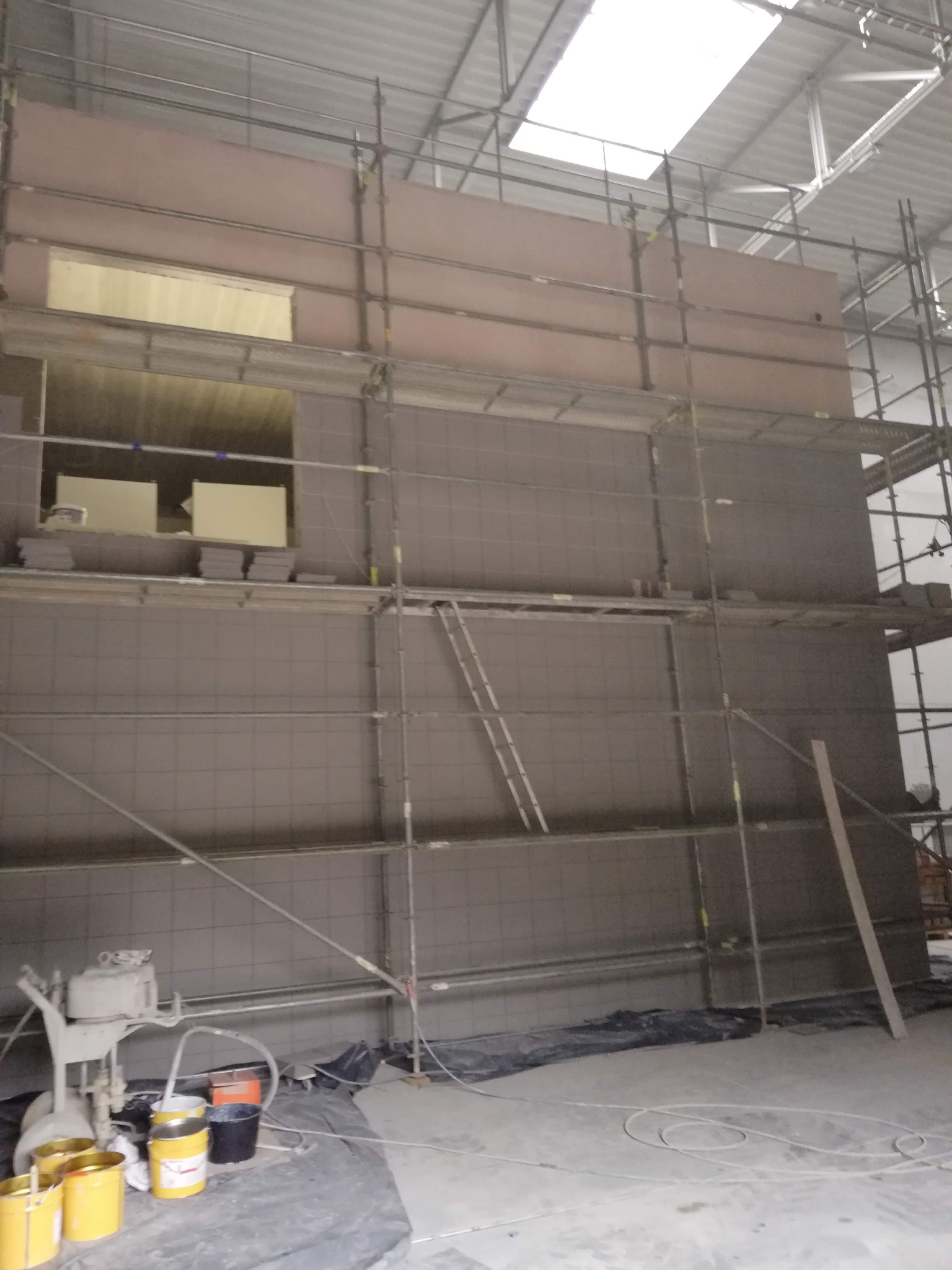 Murowana ściana w trakcie budowy, z widocznymi rusztowaniami, oknami i sprzętem do nakładania tynku.