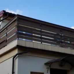 Zewnętrzny balkon z poziomą, ciemnobrązową balustradą, widok z dołu na błękitne niebo z chmurami.