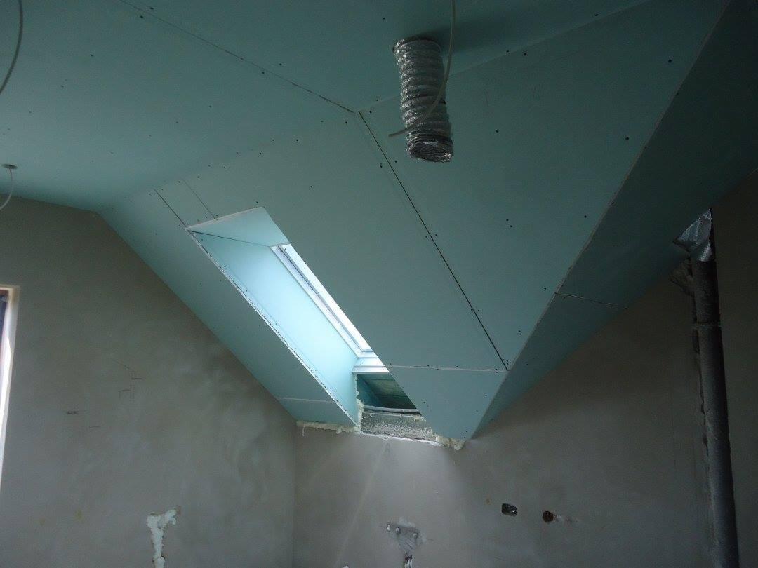 Poddasze z oknem dachowym w trakcie montażu płyt kartonowo-gipsowych, widoczne łączenia płyt i fragment instalacji wentylacyjnej.