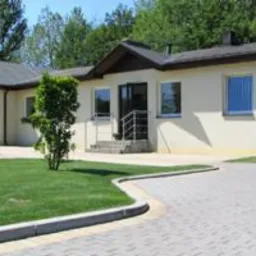 Jasny, parterowy budynek z beżową elewacją, ciemnym dachem i szarym brukowanym podjazdem. Widoczny fragment zadbanego trawnika i niewielkie krzewy. Metalowa balustrada przy wejściu.