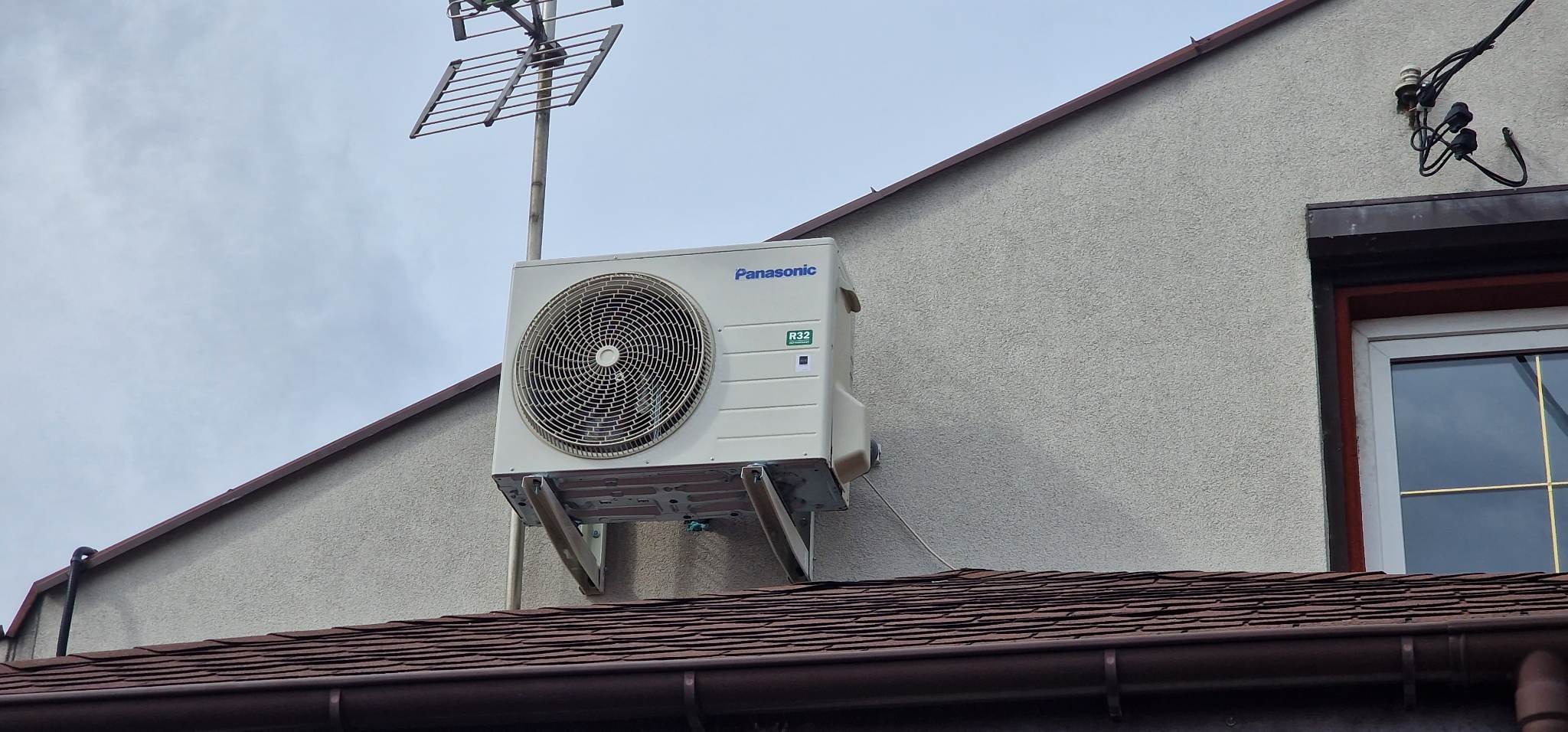Zewnętrzna jednostka klimatyzacji Panasonic R32 zamontowana na wspornikach przy ścianie domu, widoczna antena telewizyjna na dachu i okno z brązową ramą.