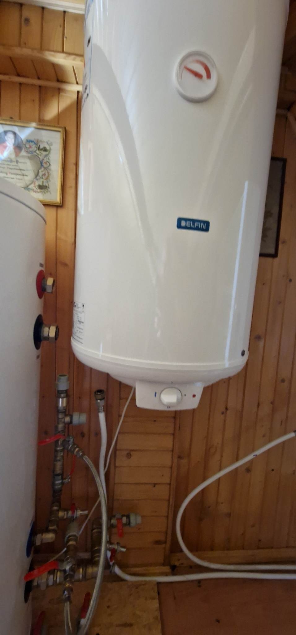 Biały, cylindryczny zbiornik na wodę marki Delfin, zamontowany na drewnianej ścianie z widocznymi rurami i zaworami, z termometrem wskazującym niską temperaturę.
