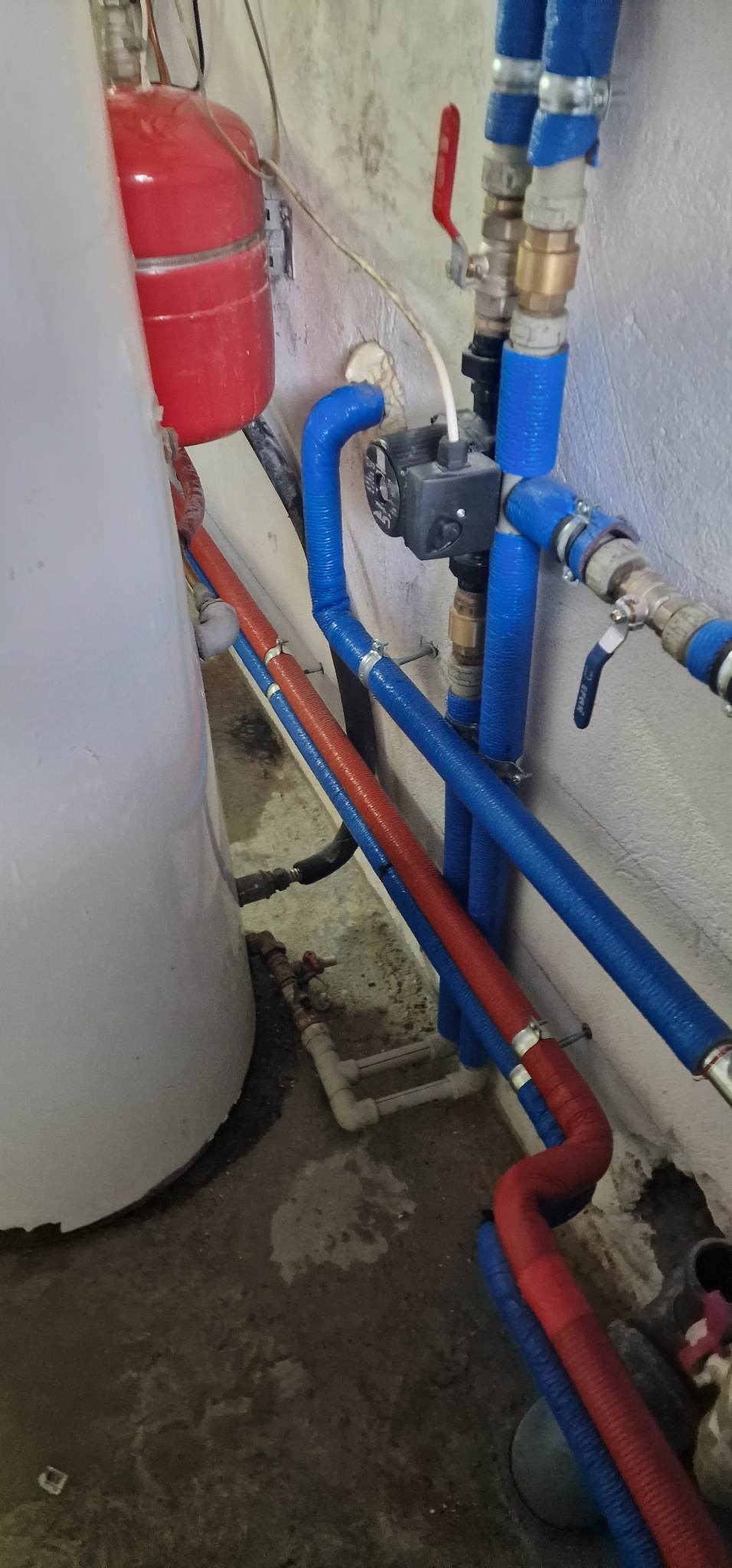 Instalacja grzewcza z czerwonym zbiornikiem wyrównawczym i rurami w izolacji termicznej, widoczna pompa obiegowa i zawory na tle surowej ściany i posadzki.
