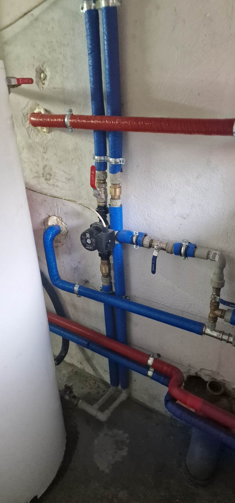 Instalacja rur w kolorze niebieskim i czerwonym, zabezpieczonych izolacją, z widoczną pompą obiegową i zaworami, zamontowana na szarej, surowej ścianie.