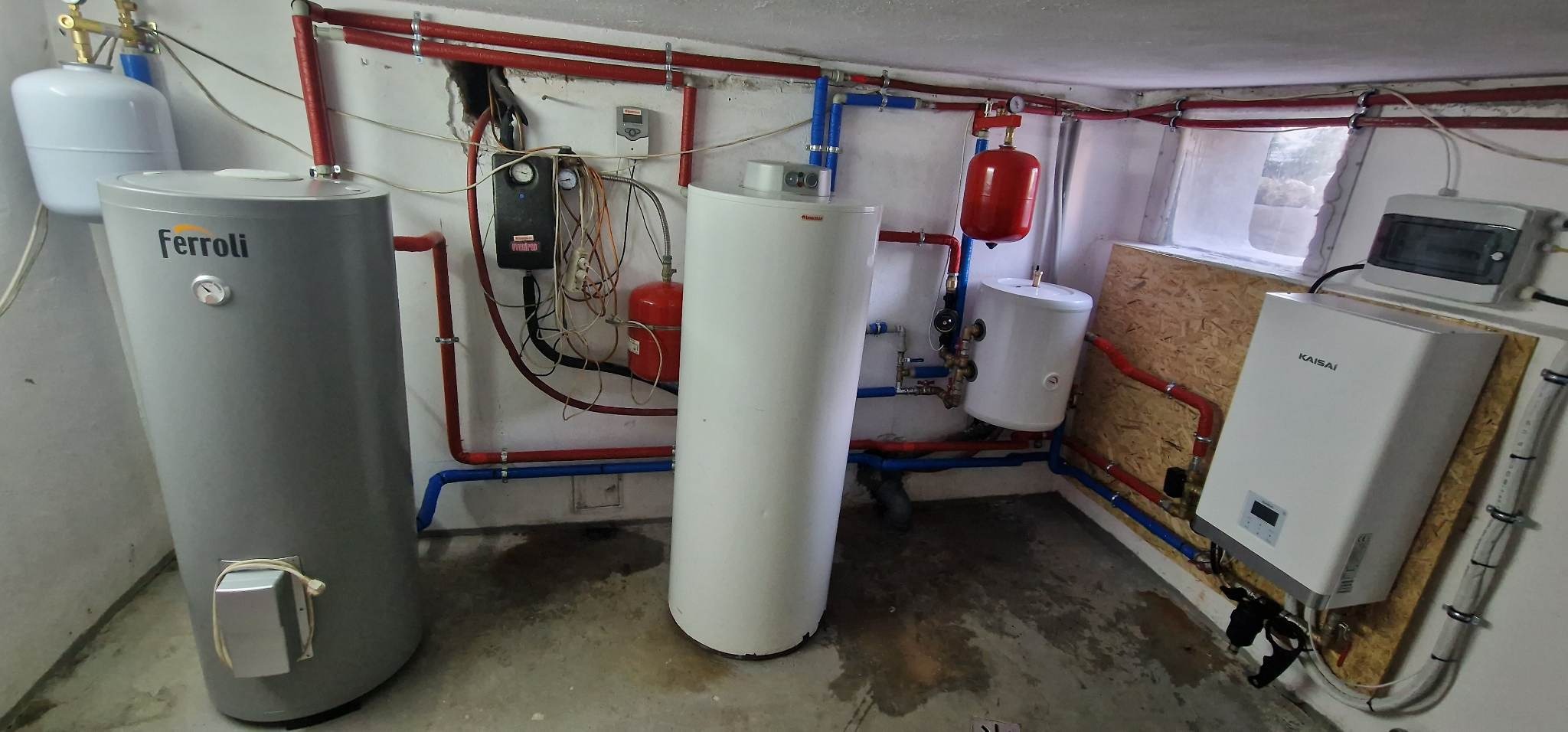 Instalacja grzewcza w piwnicy z szarym bojlerem Ferolli, białym bojlerem, czerwonym zbiornikiem wyrównawczym, czarną skrzynką sterowniczą i białą jednostką Kaisai zamontowaną na płycie OSB.