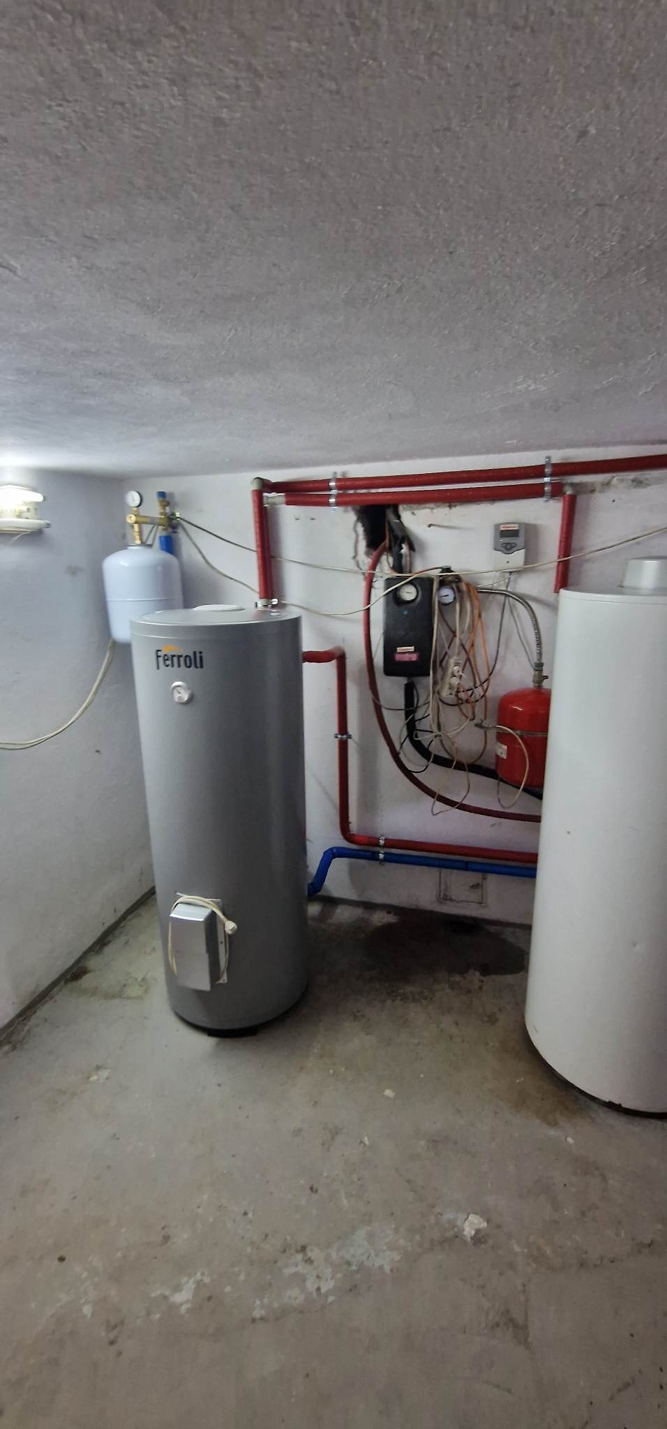 Szary, cylindryczny zbiornik Ferroli z podłączonymi rurami i kablem elektrycznym, obok biały zbiornik, na tle ściany z instalacją hydrauliczną i elektryczną w piwnicy z surowym betonowym sufitem...